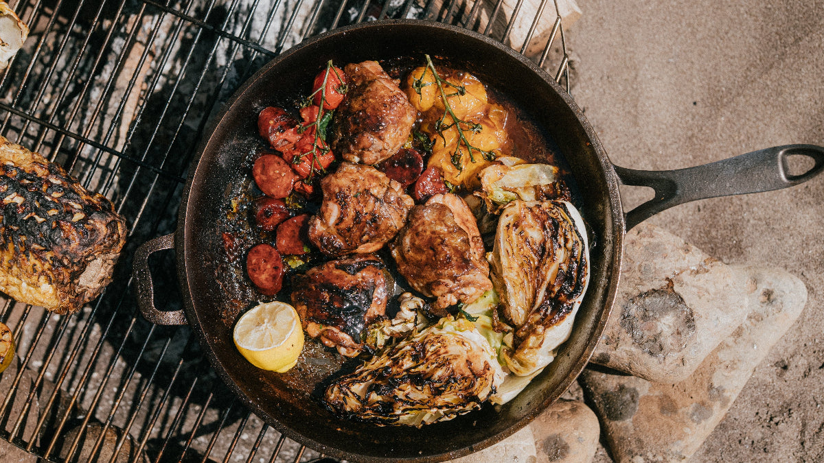 Hornbeam No12 cast iron skillet containing chicken thighs, hispi cabbage, chorizo and Isle of whight cherry tomatoes over a beach barbeque 